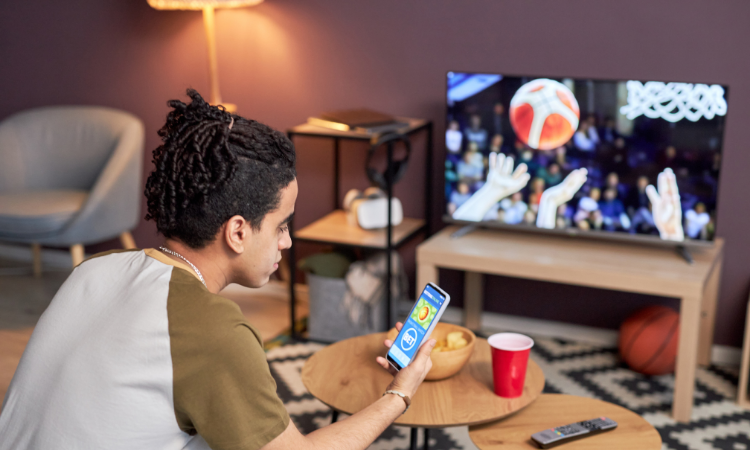 A young man sitting on a couch looking on his phone with a sports game playing on the tv in the background