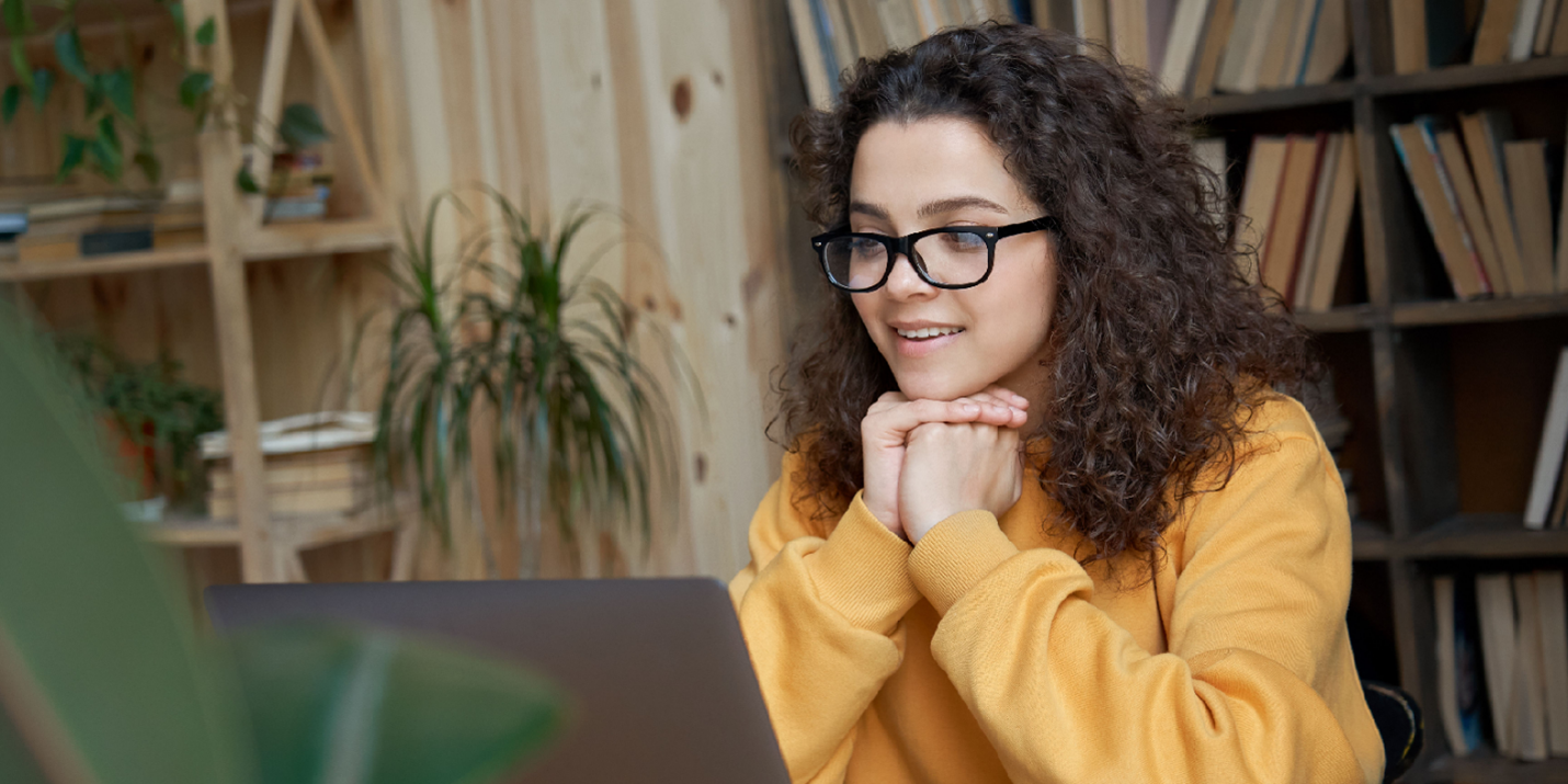 Woman with curly hair and glasses in a yellow sweater smiling thoughtfully at a laptop, with bookshelves and plants.